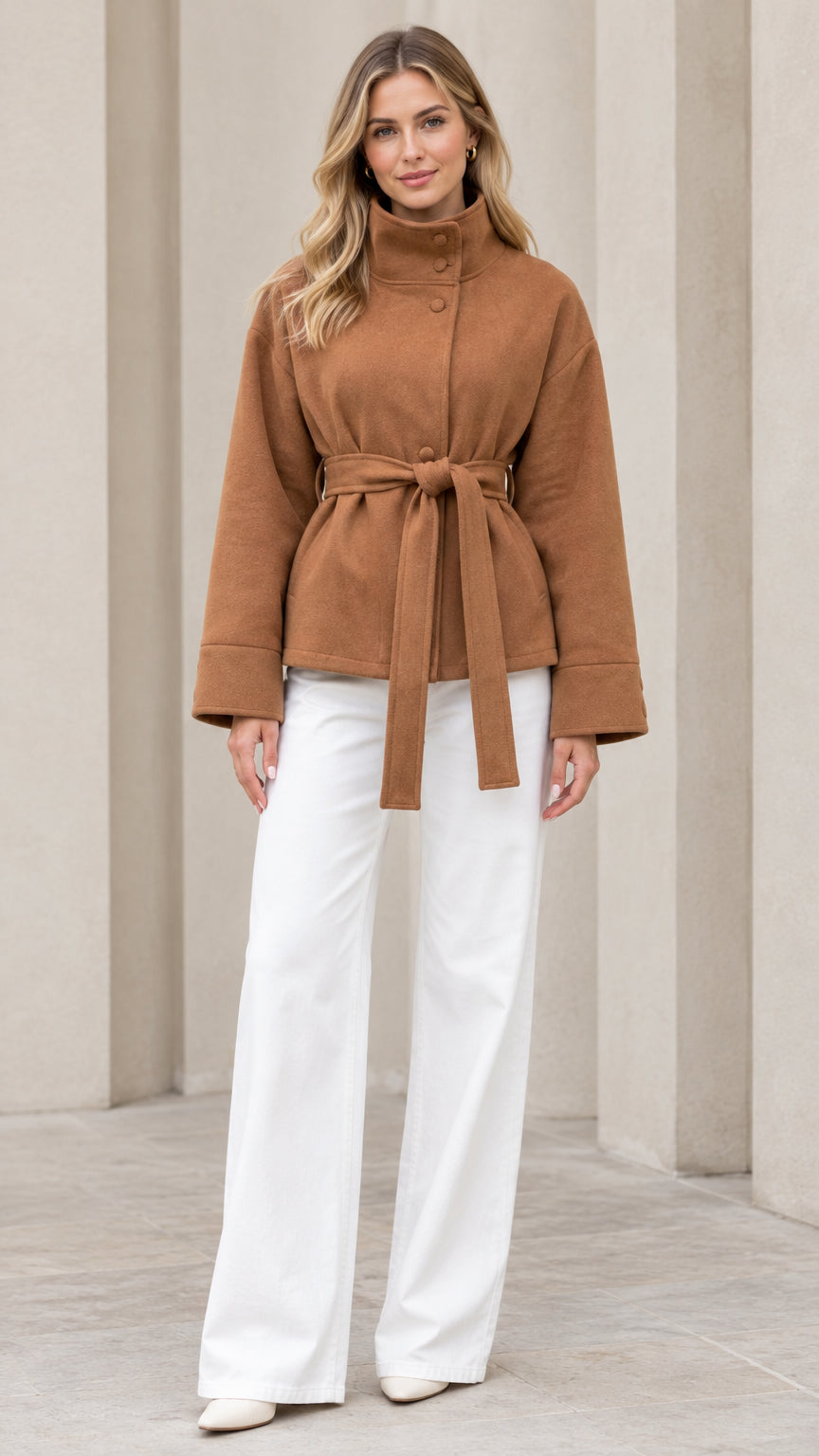 Front view of model wearing camel stand collar belted short coat with white trousers