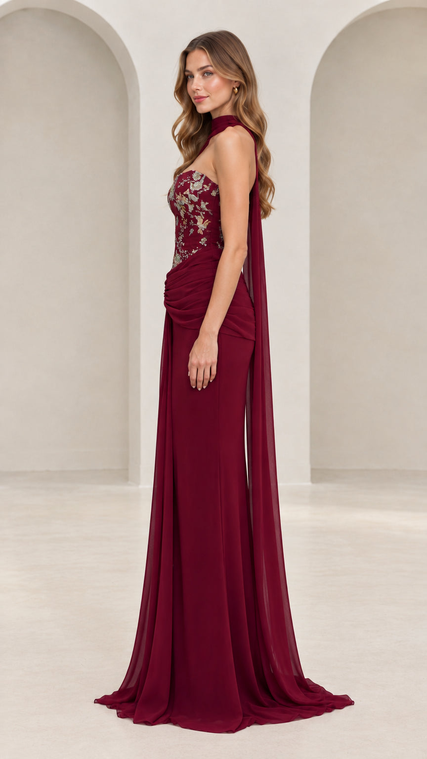 Side profile of burgundy asymmetric cape formal gown with floral embroidered bodice