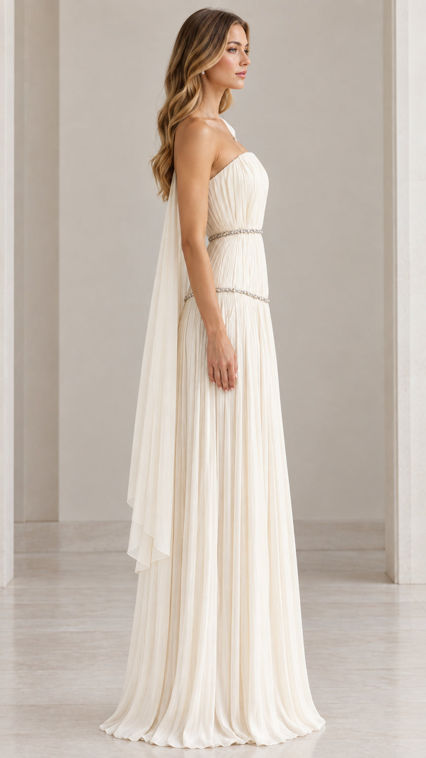 Side profile of model in ivory strapless pleated maxi gown with flowing back drape