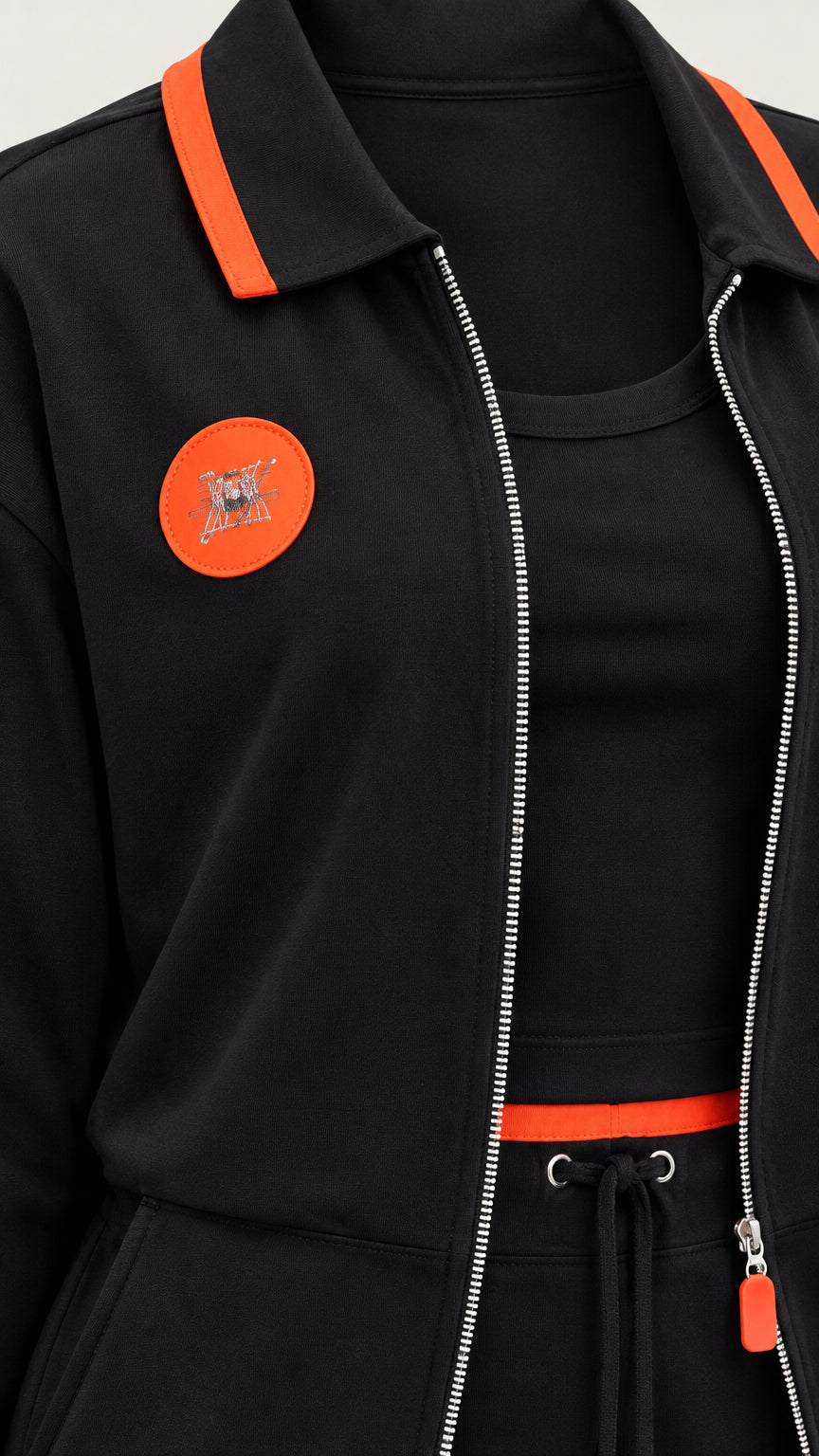 Close-up detail of black jacket collar, zipper, orange patch, and drawstring waist